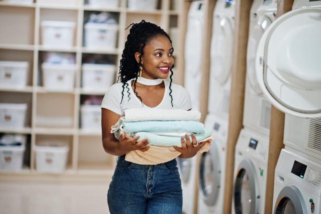 A woman holding folded laundry stands in front of washing machines at a laundromat, smiling and looking to the side.