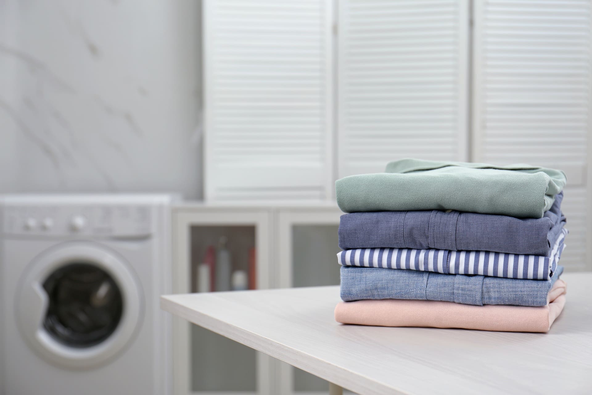 A neatly folded stack of clothes sits on a table, with a washing machine and cabinets visible in the background&mdash;showcasing the quality you can expect from a local laundry service Katy TX.