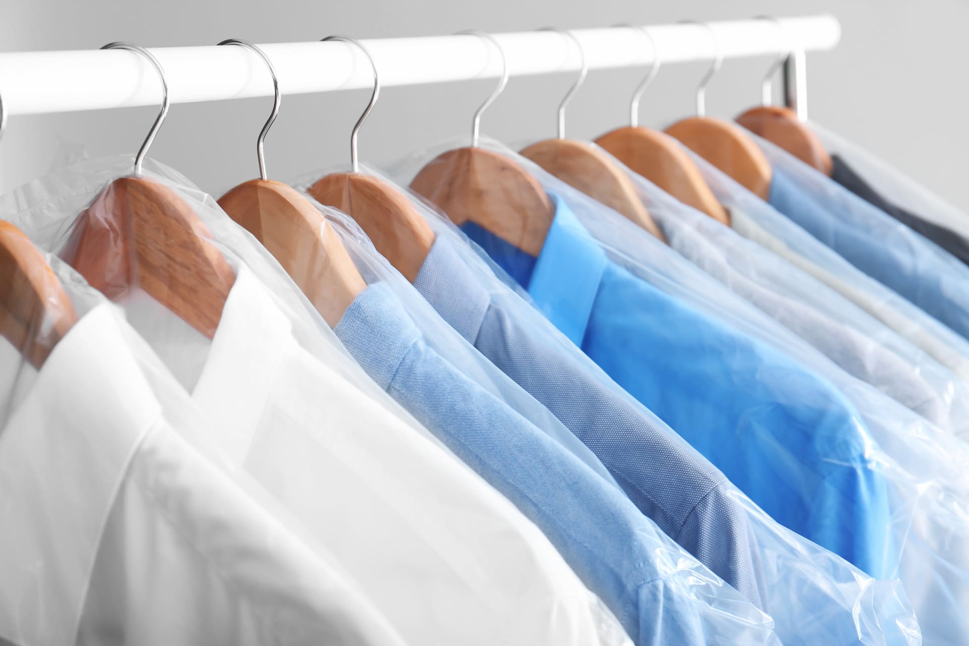 Several shirts in shades of blue and white hang on wooden hangers, each covered in plastic, on a white clothing rack&mdash;ready for pickup and delivery dry cleaning Katy.
