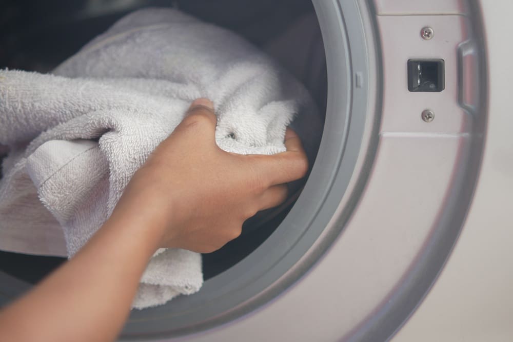 A hand placing a white towel into the open door of a front-loading washing machine.