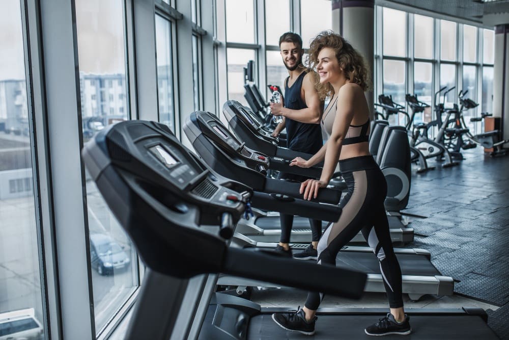Two people in athletic wear use treadmills at a modern gym with large windows and various exercise equipment in the background.