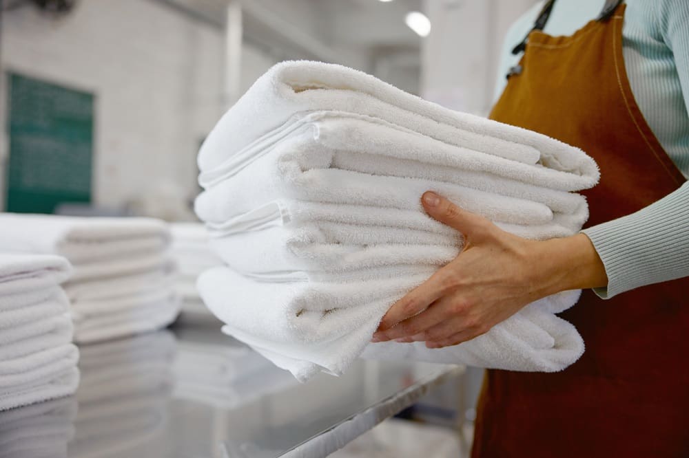 A person wearing a brown apron holds a stack of folded white towels in a laundry facility.