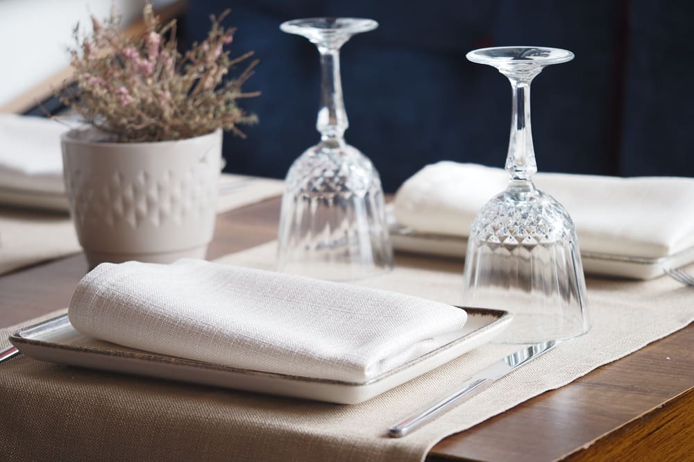 Table setting with two plates, each with a neatly folded white napkin, two upside-down wine glasses, cutlery, and a potted plant centerpiece on a wooden table.