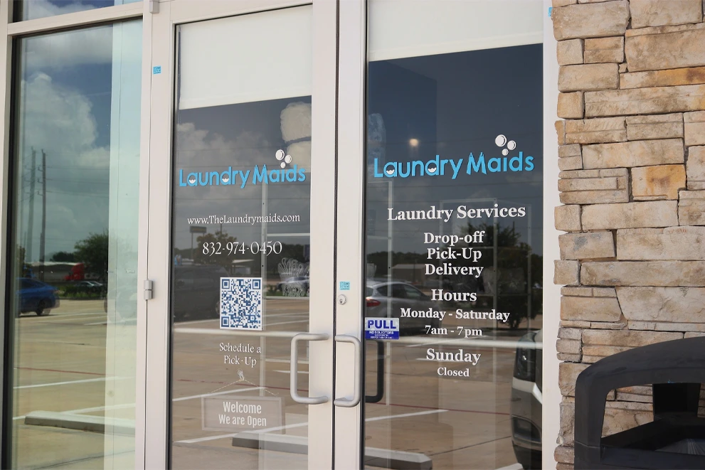 Glass doors of a laundry service business called Laundry Maids, displaying contact information, services offered, operating hours, and a pull handle.