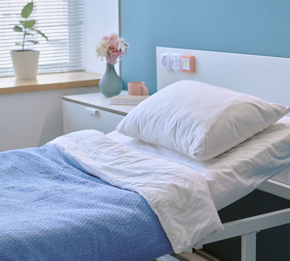 A neatly made hospital bed with white sheets and a blue blanket, beside a bedside table holding a vase of flowers, a plant, books, and a mug.
