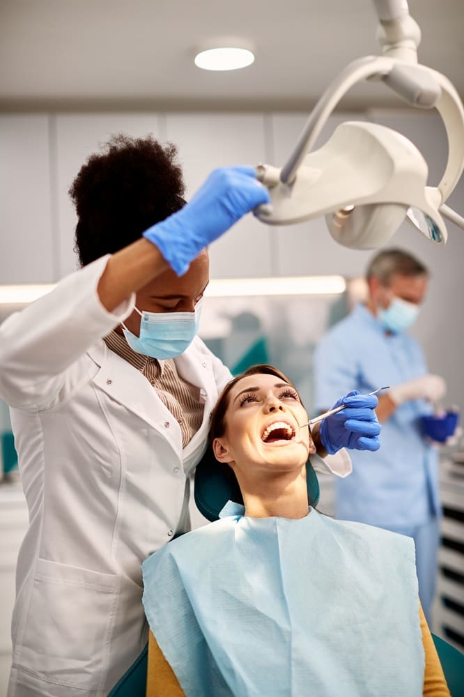 A dentist wearing gloves and a mask examines a patient’s mouth while the patient sits in a dental chair; another dental professional works in the background.