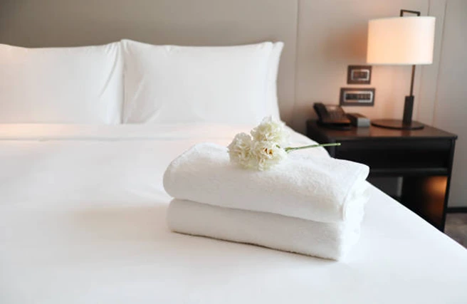 A neatly made bed with two folded white towels and white flowers placed on top, next to a dark nightstand with a lamp and telephone.