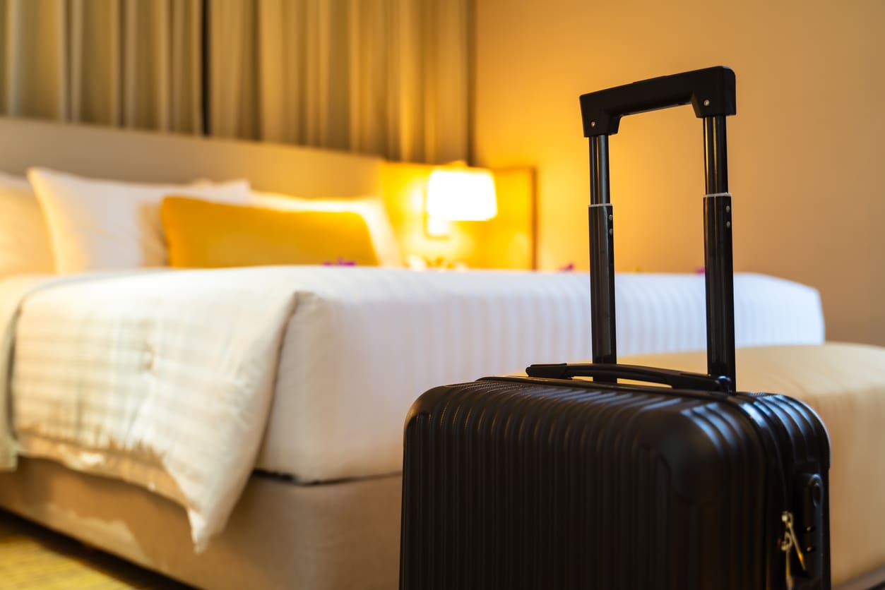 A black suitcase with an extended handle stands in front of a neatly made bed in a warmly lit hotel room.