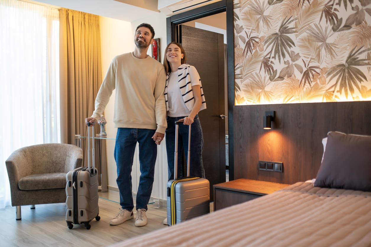 A man and woman holding hands enter a hotel room with suitcases, standing near the bed and looking around.