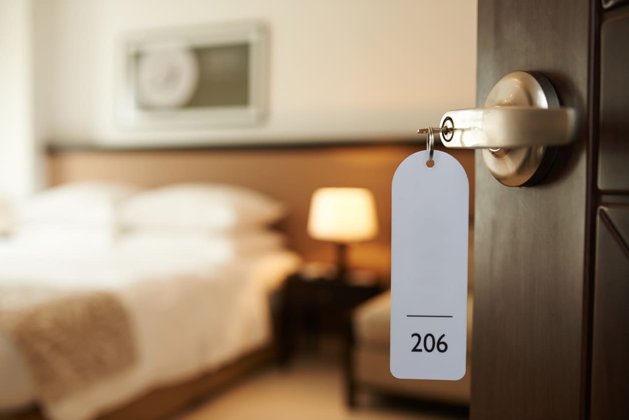 A hotel room door with a key tag labeled "206" hangs in the lock; a neatly made bed and a lit lamp are visible inside the softly lit room.
