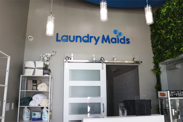 Front counter of a laundromat named "Laundry Maids" with shelves holding towels and supplies, a sliding door, and a wall with green plants.
