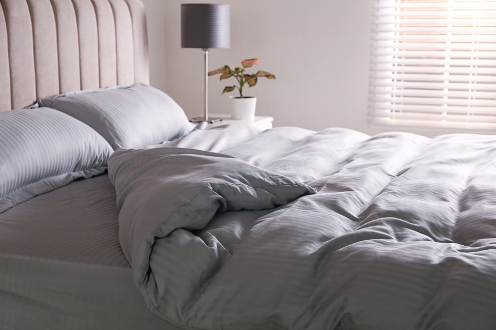 A neatly made bed with light gray striped bedding, a tufted headboard, a bedside table with a lamp, a potted plant, and a window with blinds letting in natural light.