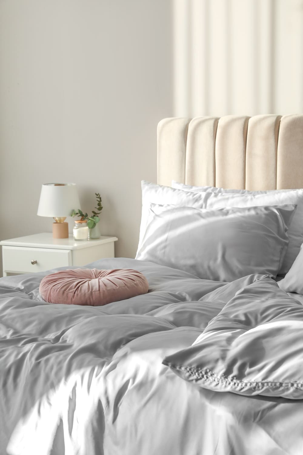 A neatly made bed with light gray bedding, a pink round pillow, and a beige upholstered headboard beside a white nightstand with a lamp and small decor items.