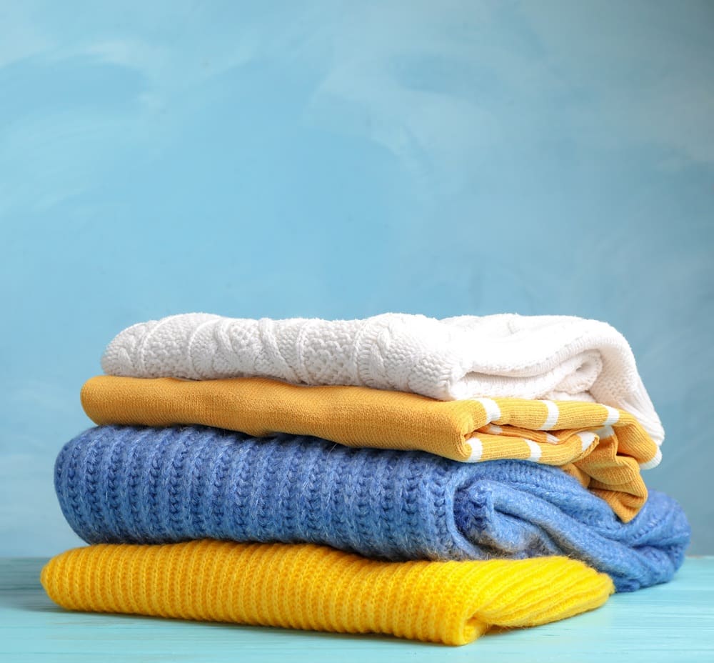 A neatly folded stack of sweaters and towels in white, yellow, and blue sits on a turquoise wooden surface against a light blue background.