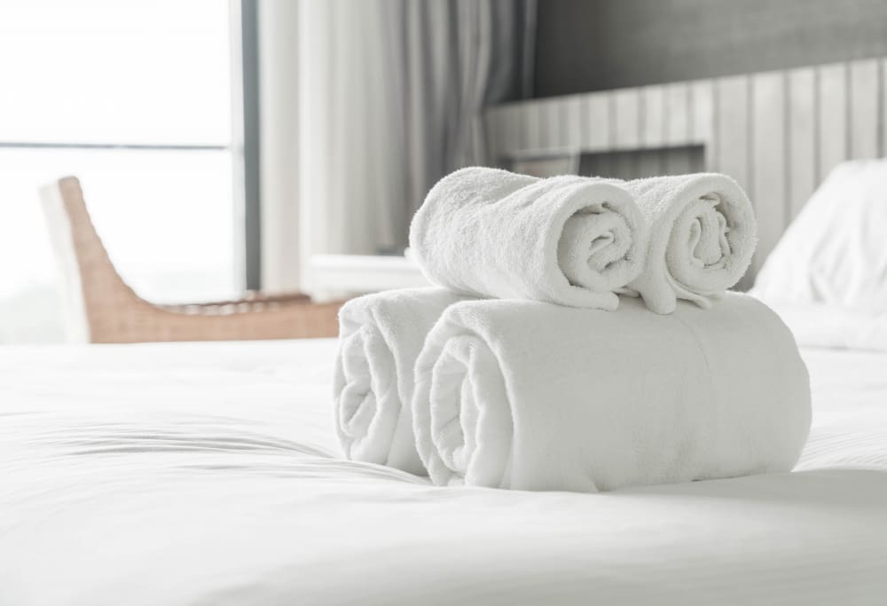 A stack of neatly rolled white towels sits on a white bed in a bright, modern bedroom with a wicker chair in the background.