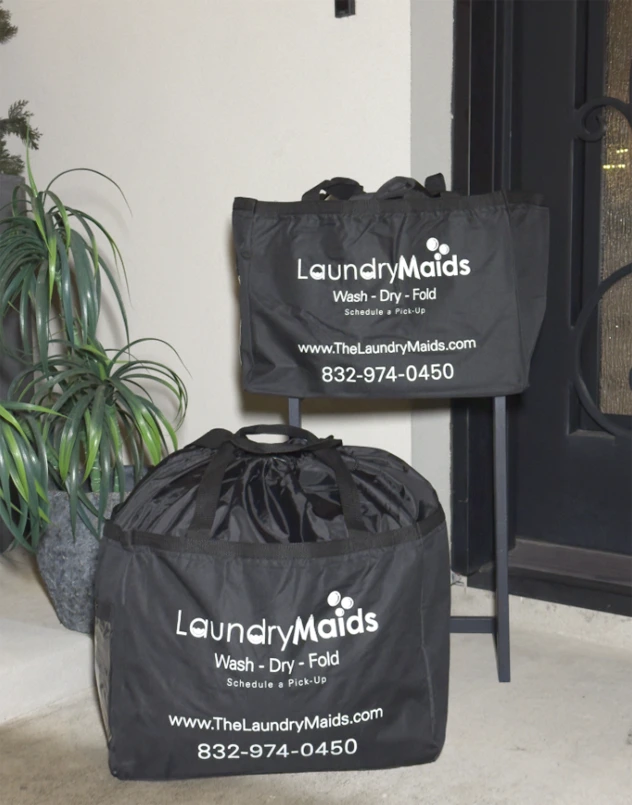 Two black laundry bags with "Laundry Maids" logo, contact number, and website are placed on a porch near a potted plant and a black door.
