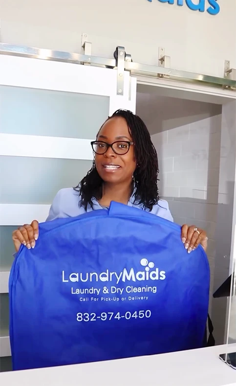 A woman stands behind a counter holding a blue LaundryMaids garment bag with the company's phone number printed on it.
