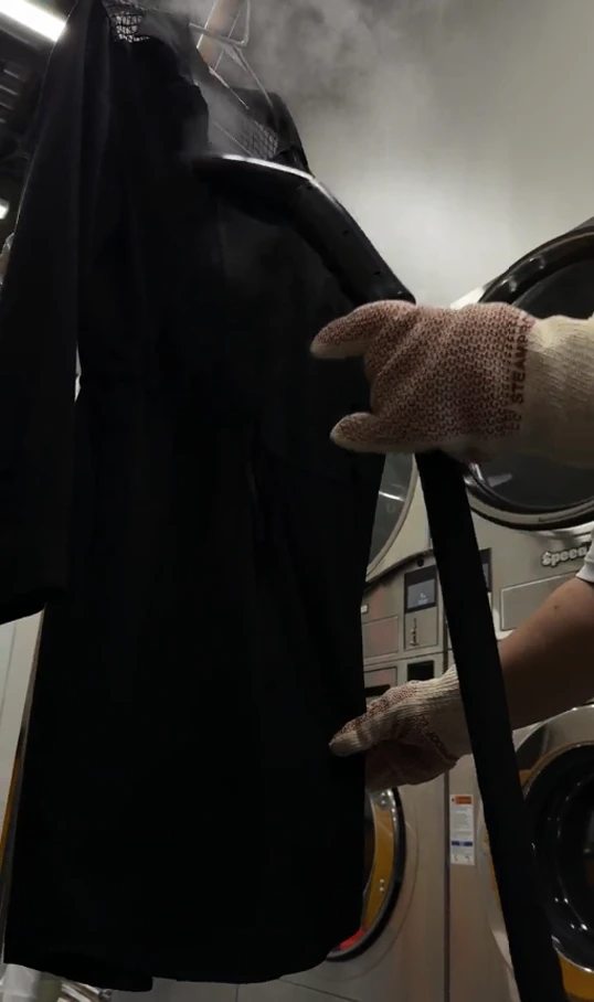 A person wearing gloves uses a steam machine to press a black garment in front of industrial washing machines in a laundry facility.