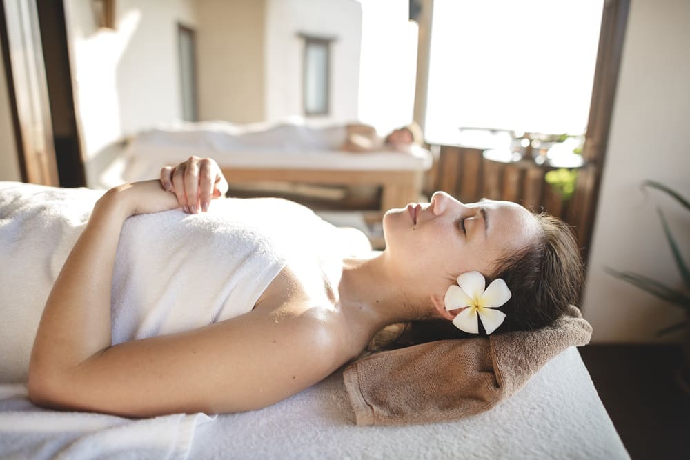A woman with a flower in her hair lies on a massage table covered with a towel, relaxing at a spa. Another person is visible in the background on a separate table.