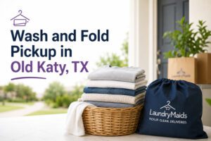 A basket of folded clothes and a LaundryMaids bag on a table, with text: "Wash and Fold Pickup in Old Katy, TX." A house entrance and delivery box are in the background.